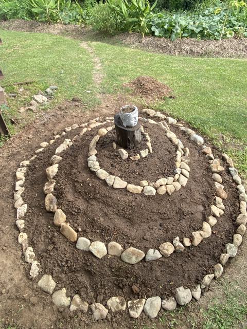 A stone herb spiral laid out on bare soil with a carved head on a stump at the centre