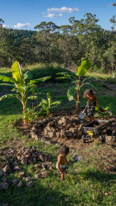 Joel shovelling compost into a rock-edged banana circle while a small child helps with a shovel