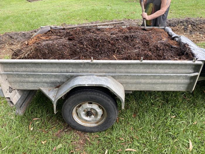 A galvanised trailer loaded with mulch being unloaded with a shovel