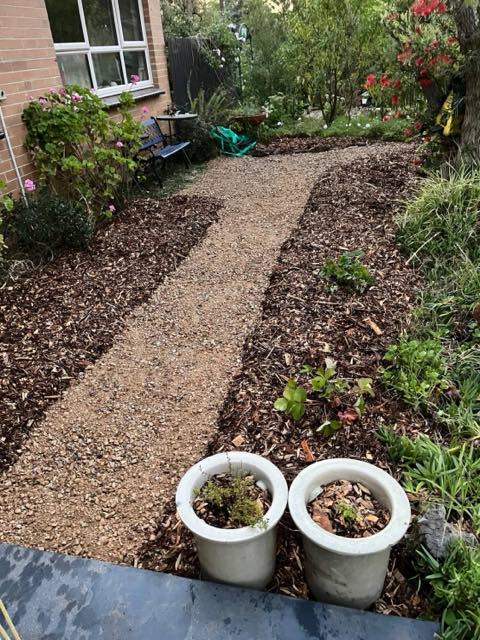 A finished gravel path with mulched and planted beds on both sides, brick house and bench beyond