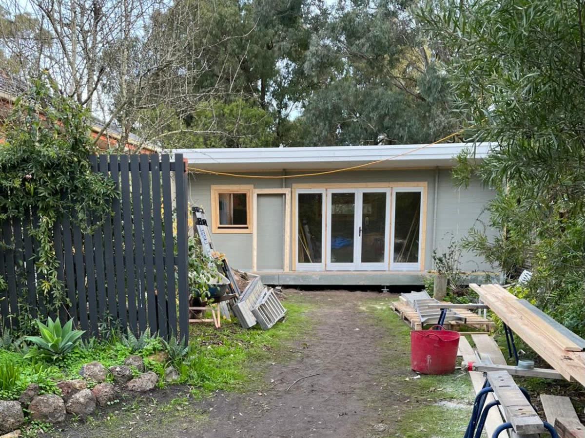 A bare dirt path beside a dark fence leading to a newly built studio, with building materials and sawhorses around