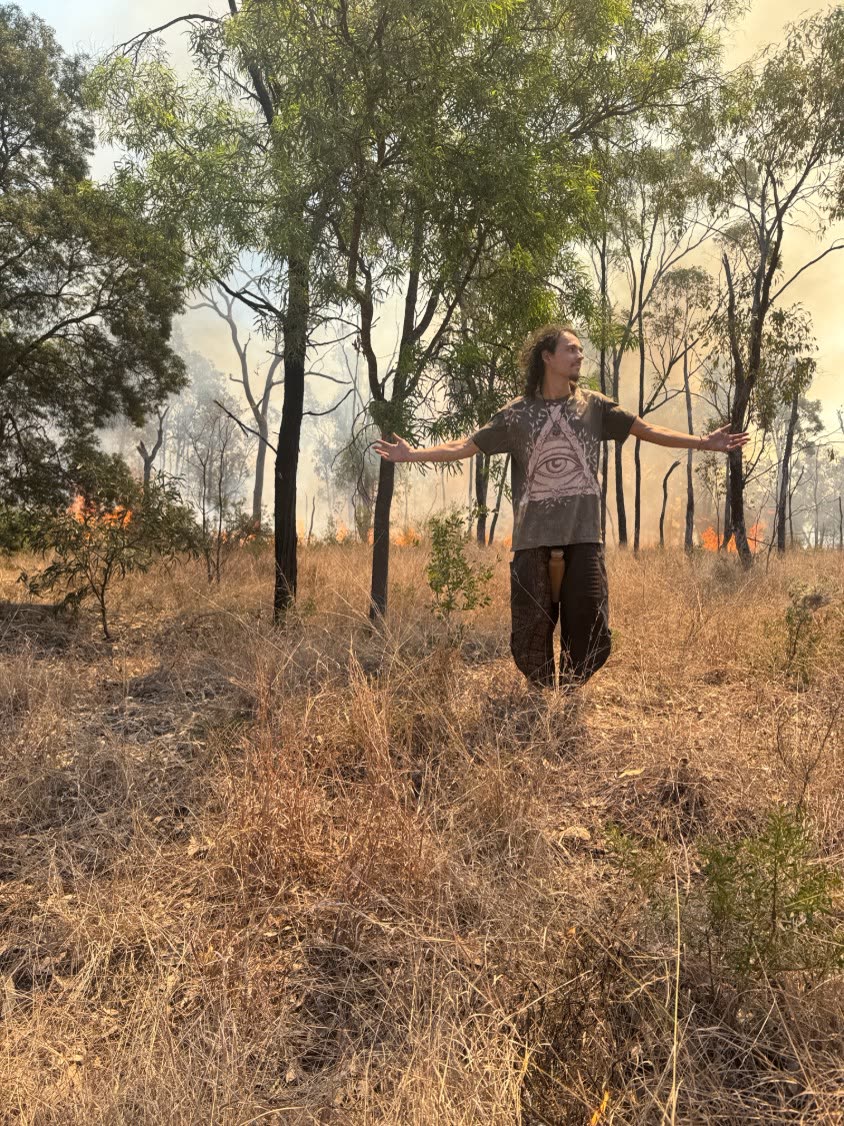 A person standing with arms out in dry grassland, a low fire and smoke moving through the trees behind