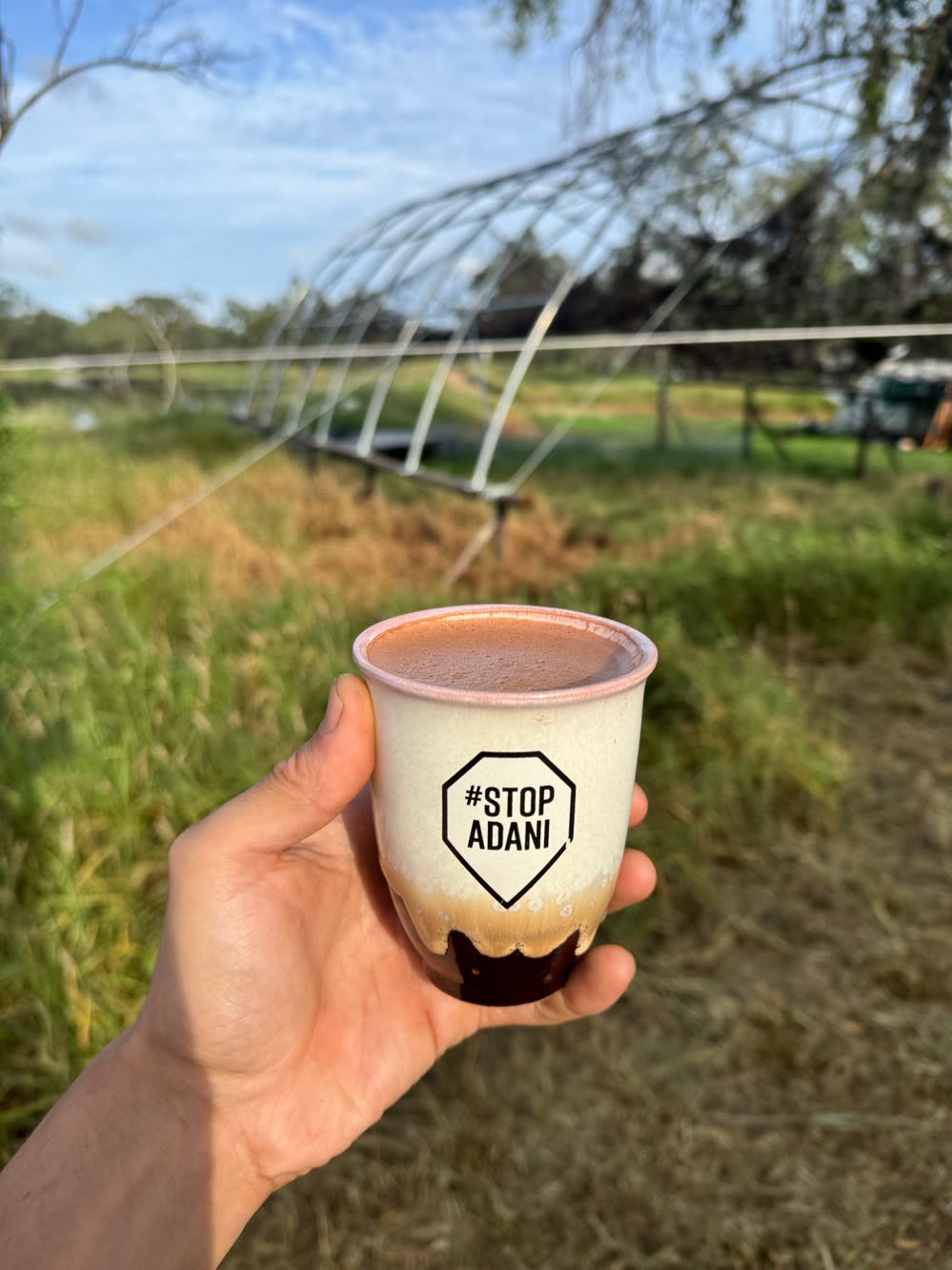 A hand holding a ceramic cup with a Stop Adani sticker, a polytunnel frame and grass behind it