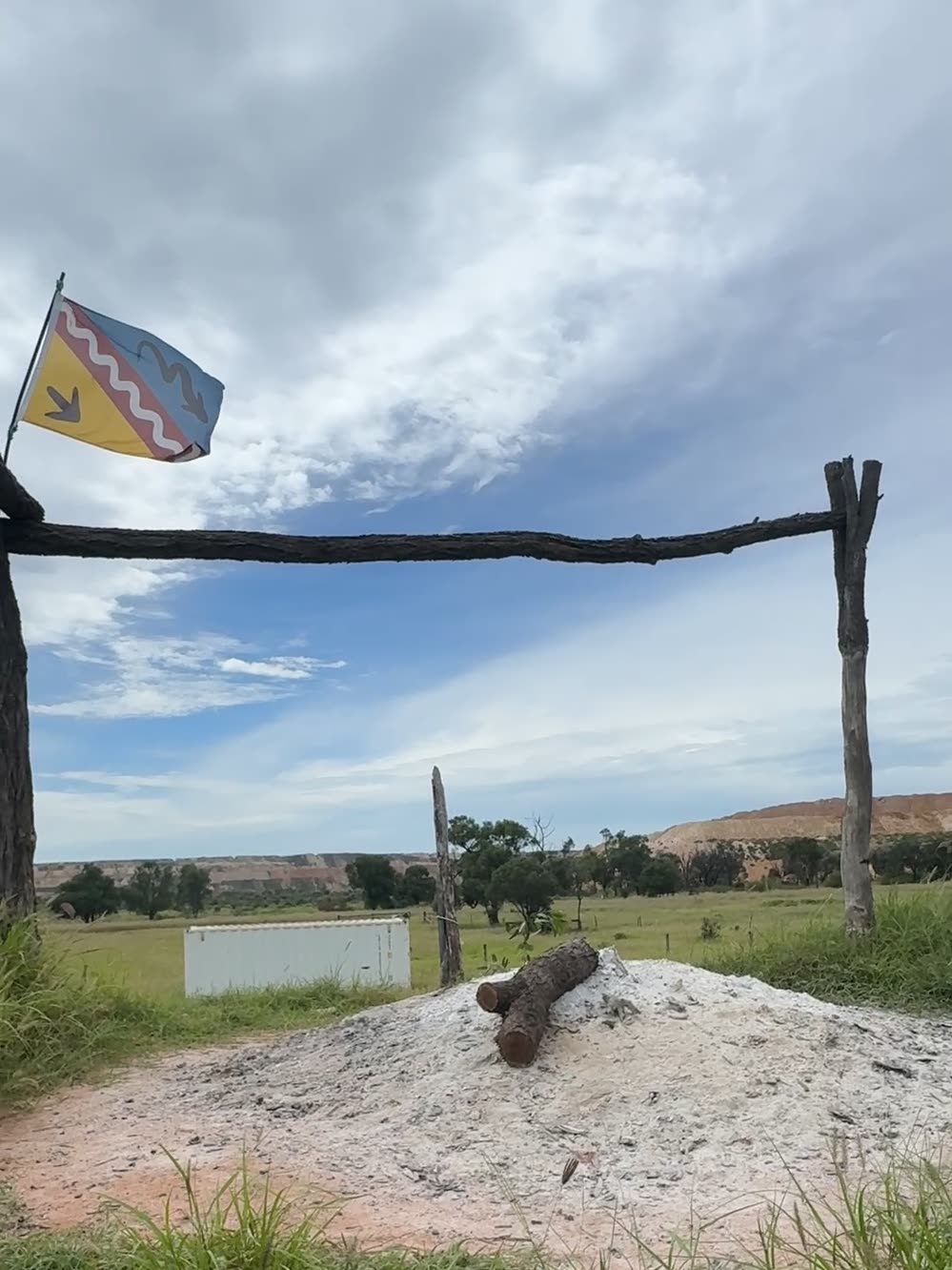 A sacred fire beneath a timber frame flying a flag, its coals banked under a mound of pale ash, mine workings on the horizon behind